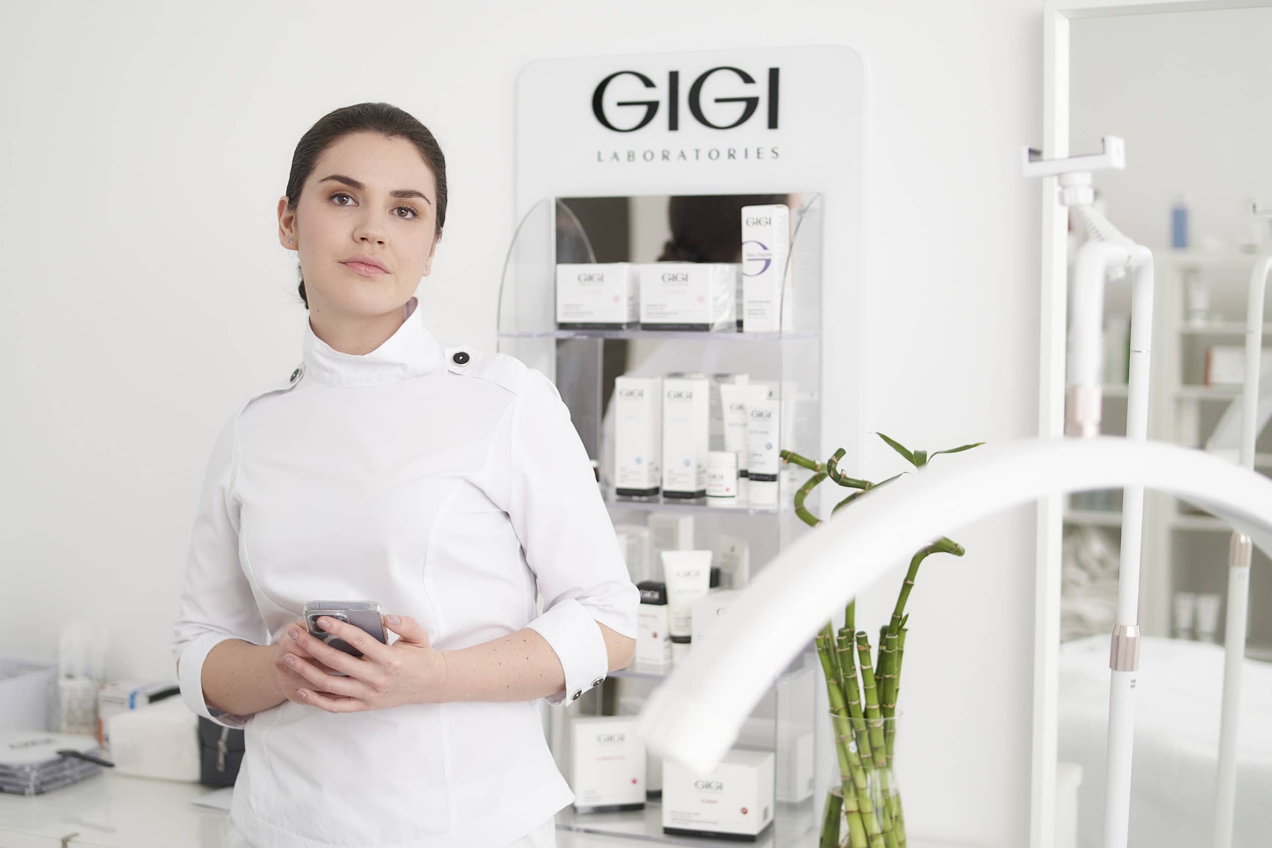 Professional beautician in white uniform standing in modern beauty salon with GIGI skincare products display