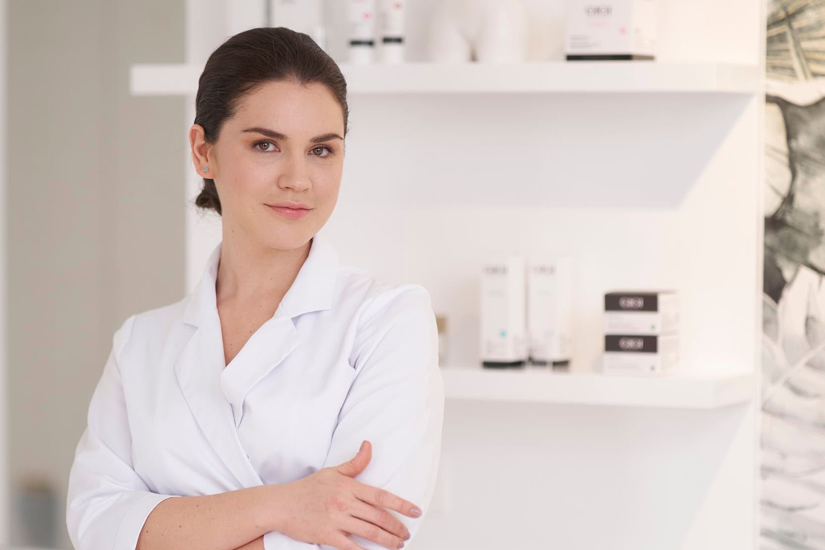 Beauty professional Regina Pronina in white robe standing with arms crossed in modern white salon