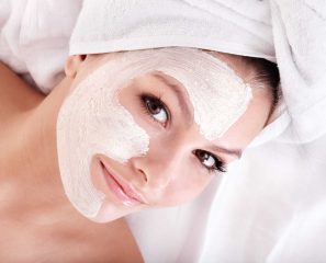 Woman with white facial mask treatment looking at camera, wearing head wrap during professional skincare therapy session.